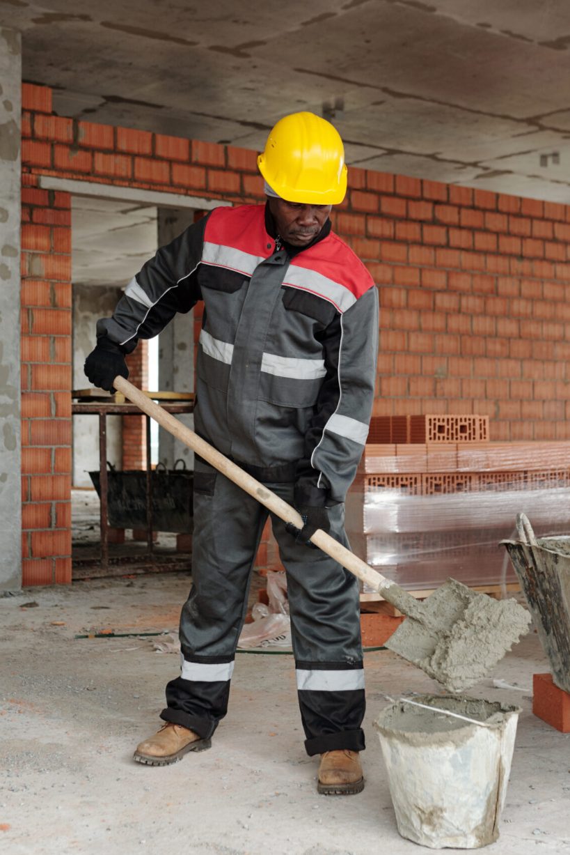 Mature black man in hardhat and workwear putting concrete into bucket while standing by large metallic basin against stack of bricks